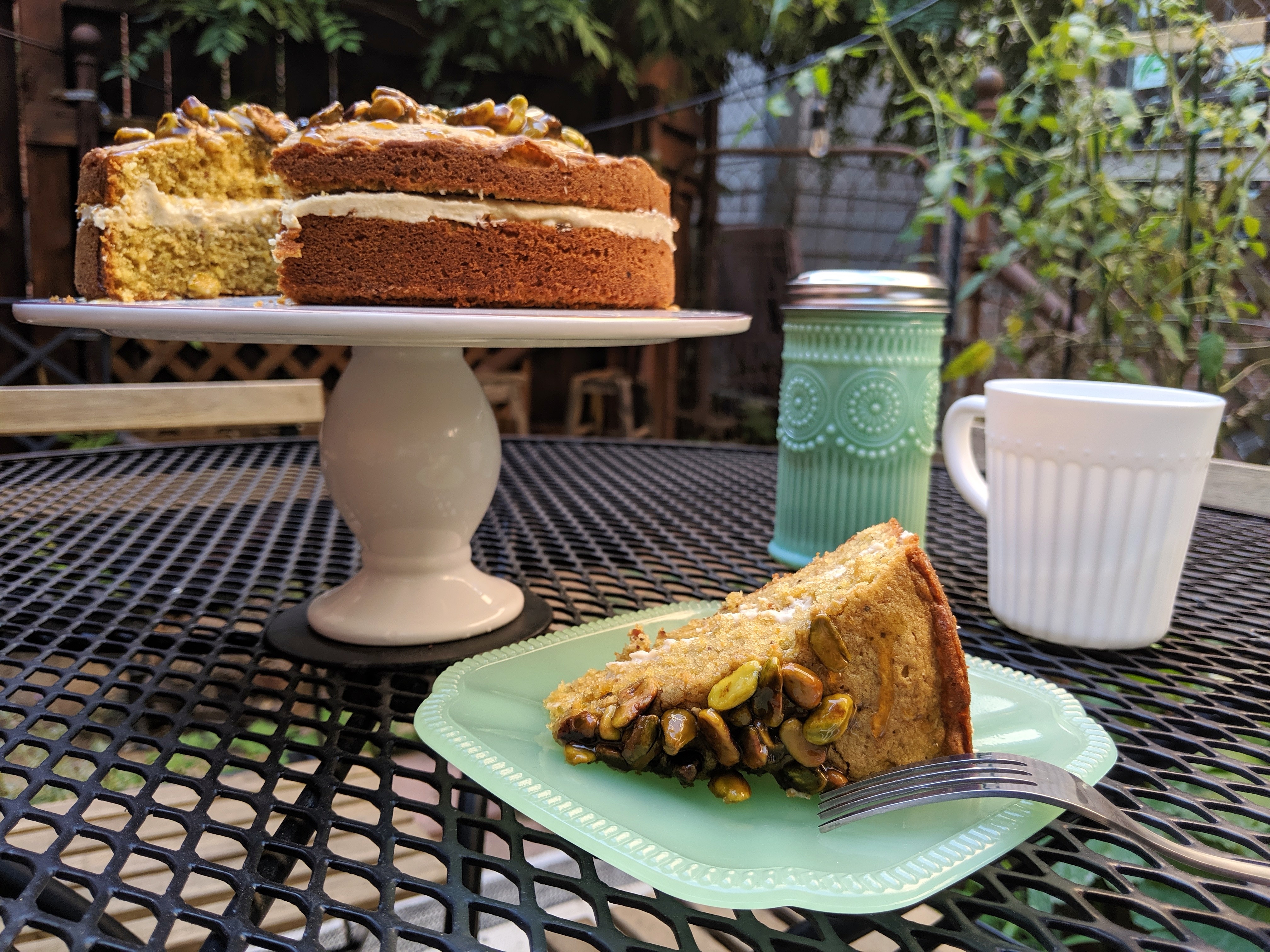 Slice of honey pistachio cake on a plate with the full cake and a mug in the background
