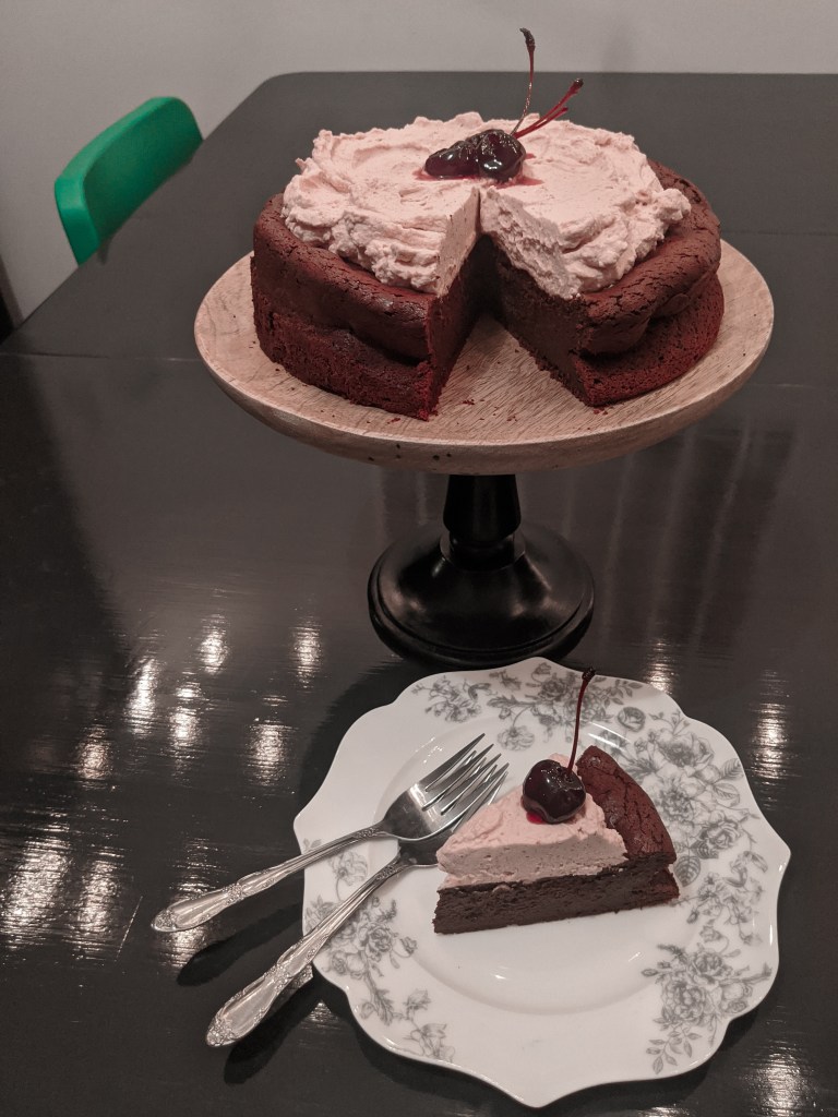A chocolate cake with black cherry whipped cream, with a slice cut out and sitting on a plate with two forks.