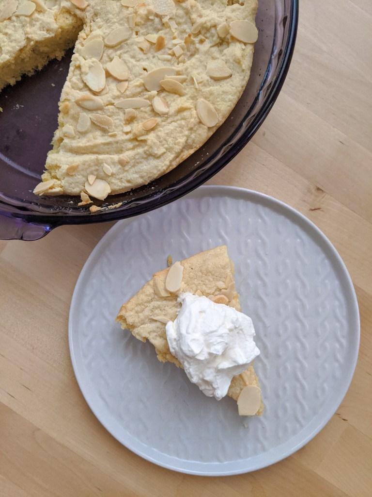 A slice of almond cake with whipped cream sitting on a plate next to the full cake.