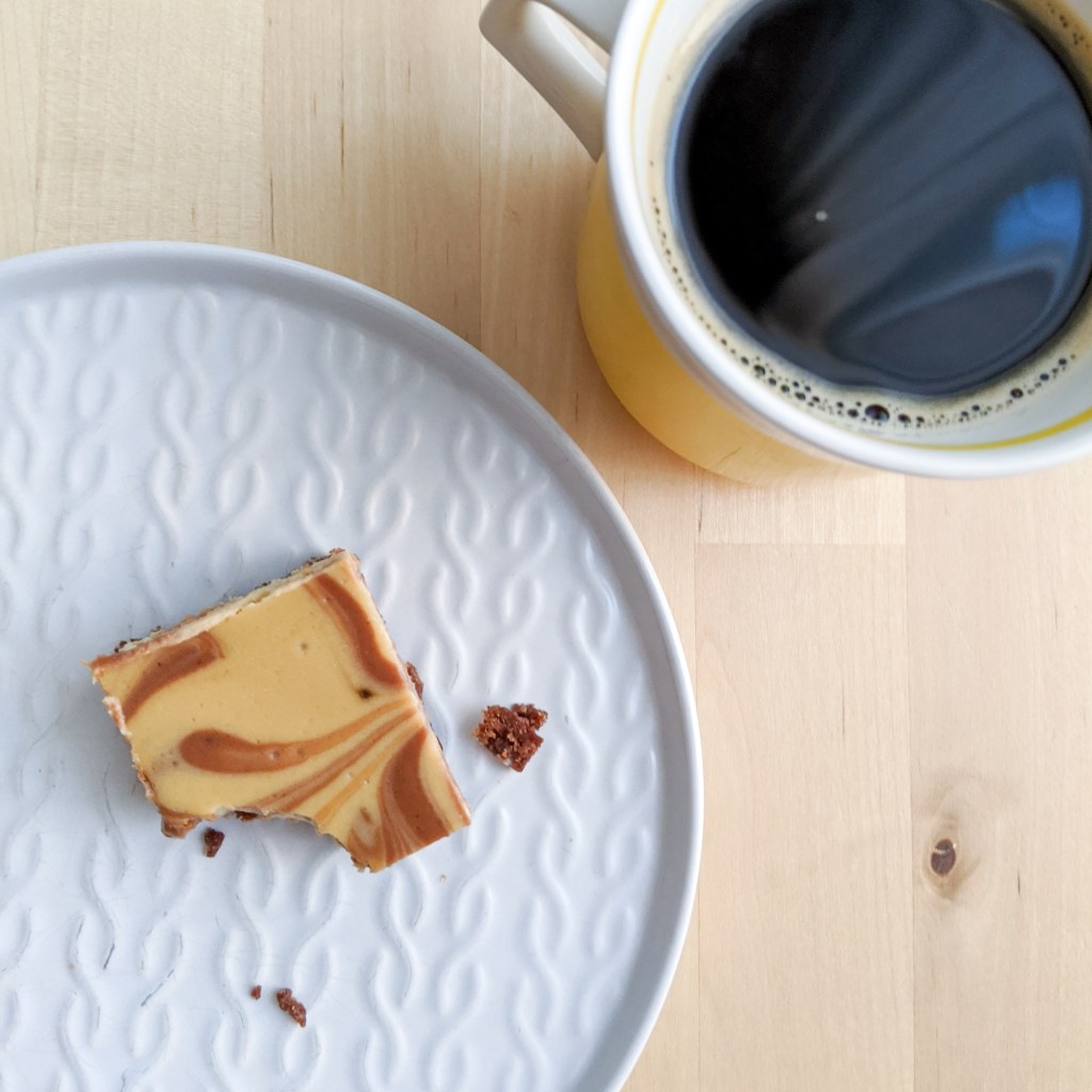 A square of marbled chocolate/coffee cheesecake with a bite missing, next to a cup of coffee