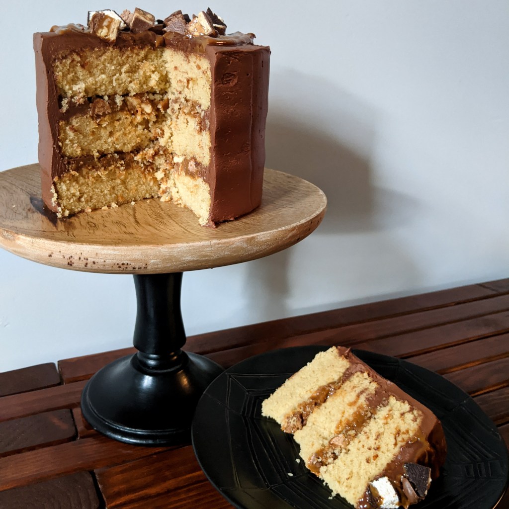 A small three-tier caramel cake with dulce de leche and candy chunks in between the layers and chocolate frosting on the outside, all resting on a wooden cake stand. A slice of the cake is on a plate next to it.