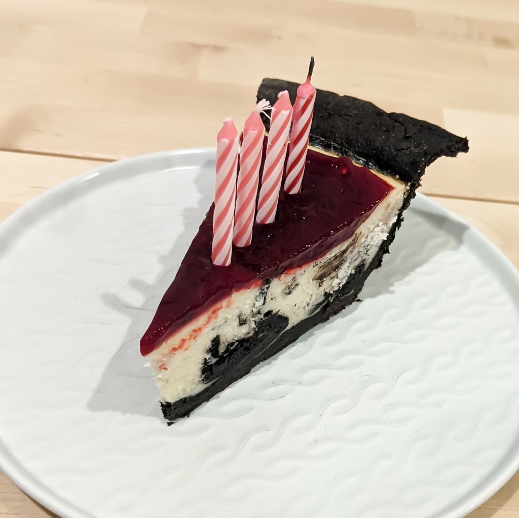 A slice of chocolate-swirled cheesecake with chocolate crust, raspberry topping, and four pink birthday candles