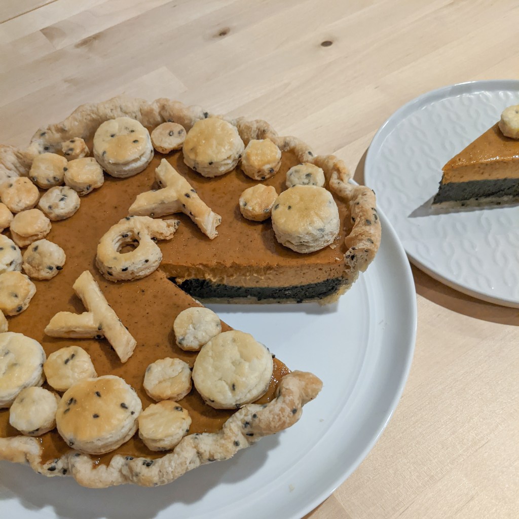 A layered pumpkin and black sesame pie with one slice cut out; the top is covered in circles of pie crust and the word "yay" spelled in crust