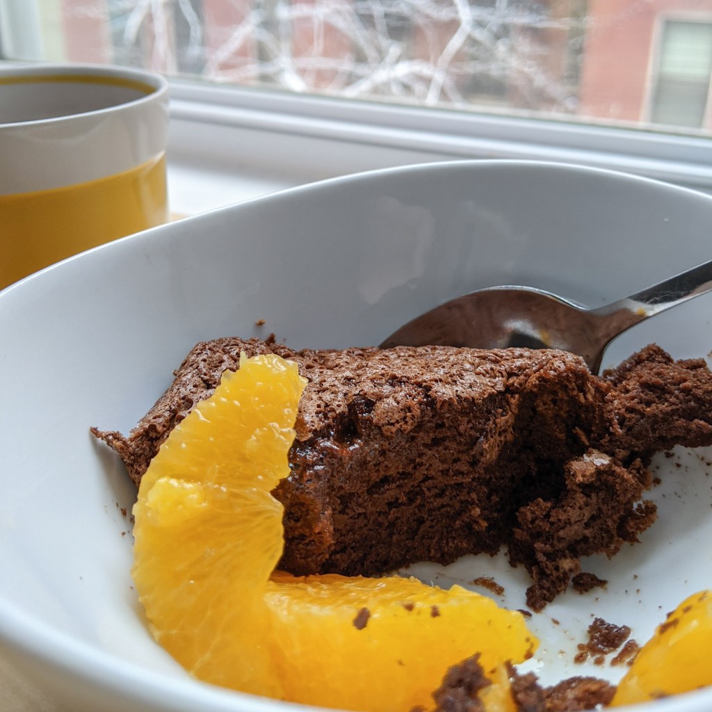 A scoop of chocolate pudding cake and some orange segments in a bowl with a mug of tea alongside.