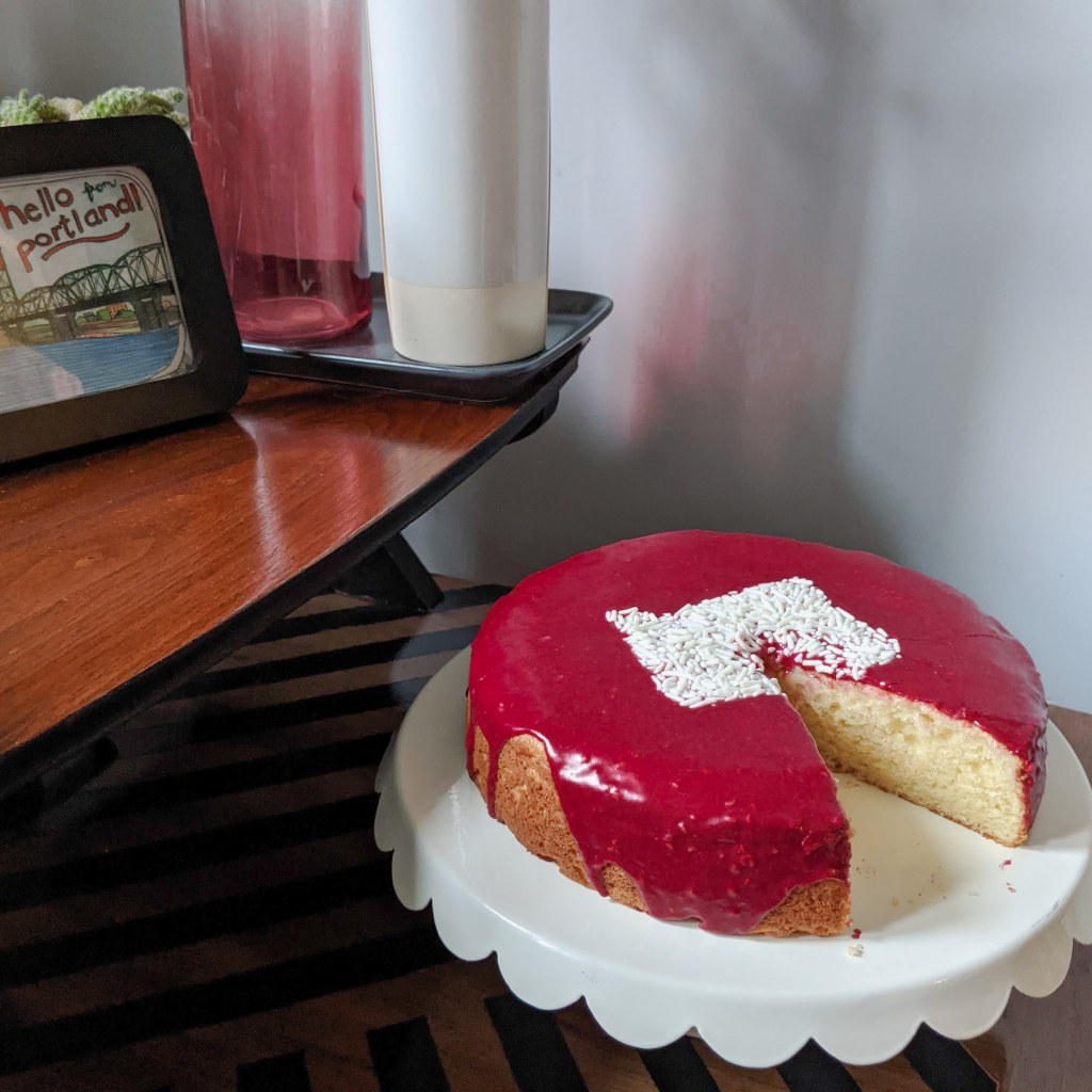 A well-decorated side table with a pink-glazed cake resting on a cake stand. The cake has the shape of the state of Oregon on the top in white sprinkles.