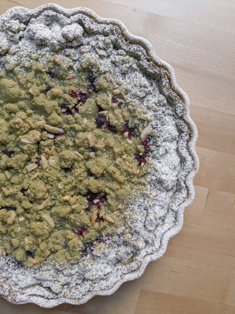 A top-down view of a green matcha streusel tart with blueberries peeking through the topping and powdered sugar around the edges