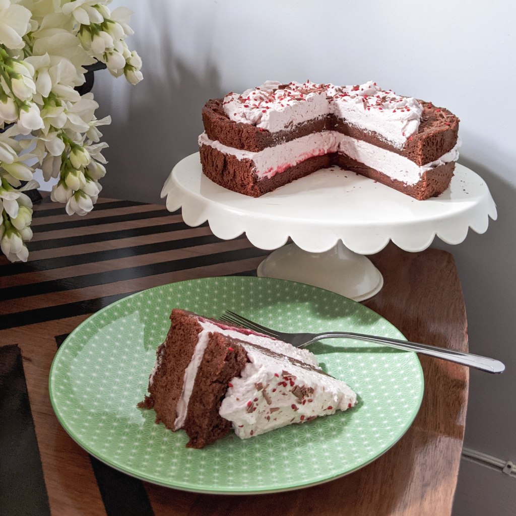 A chocolate cake with pink whipped cream on top and between the layers; a slice has been cut out and is sitting on a green plate next to the cake stand.