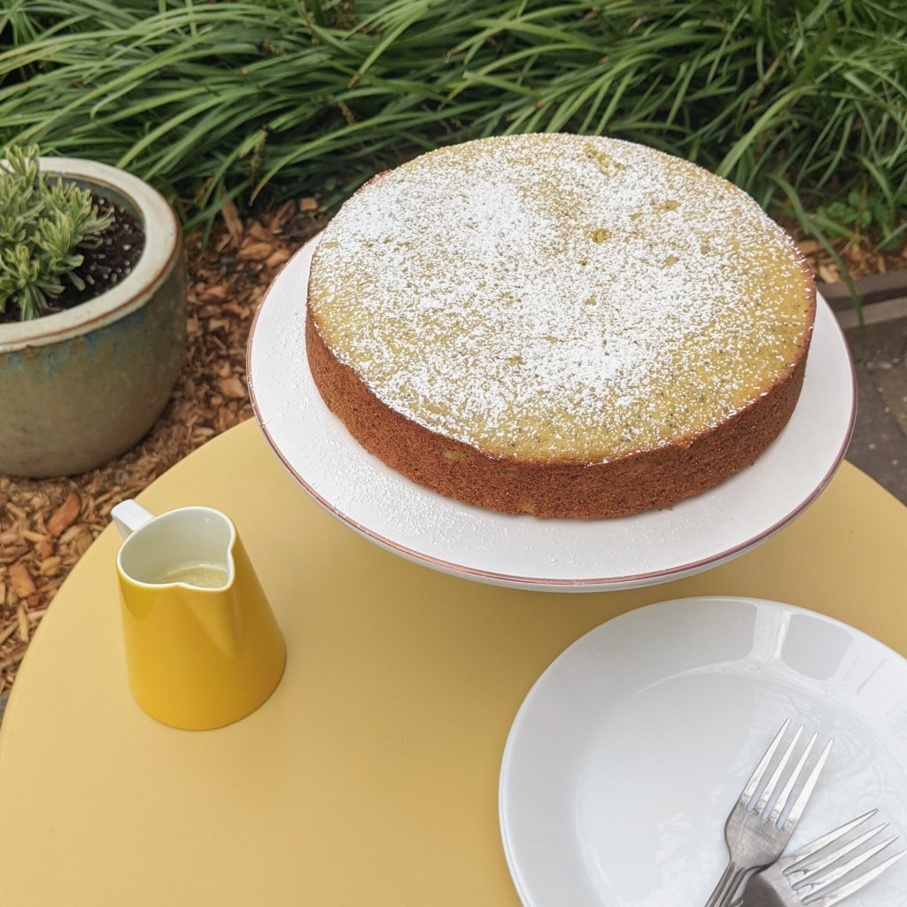 A round lemon-poppyseed cake on a cake stand, with a small pitcher of glaze and a stack of plates next to it