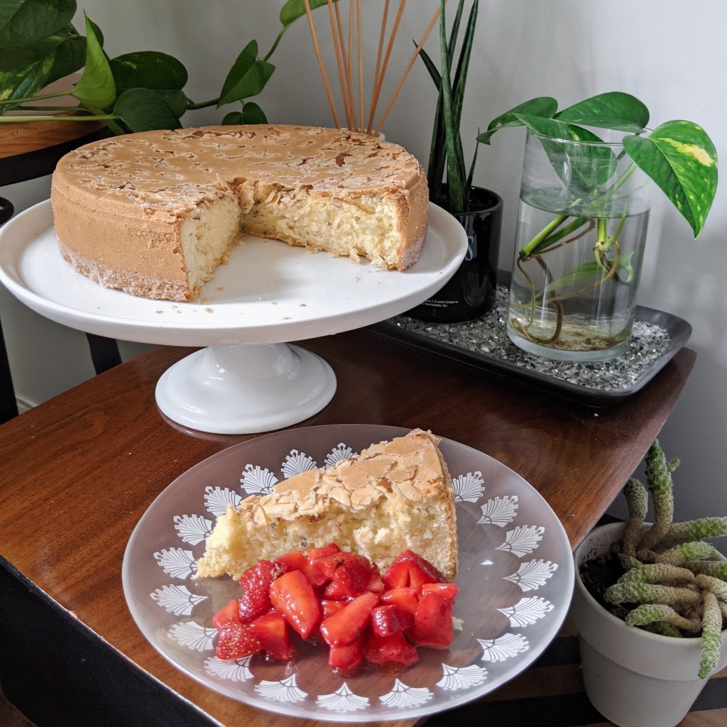 An almond-topped cake on a stand, with a slice cut out. The slice is on a plate with strawberries next to the stand.