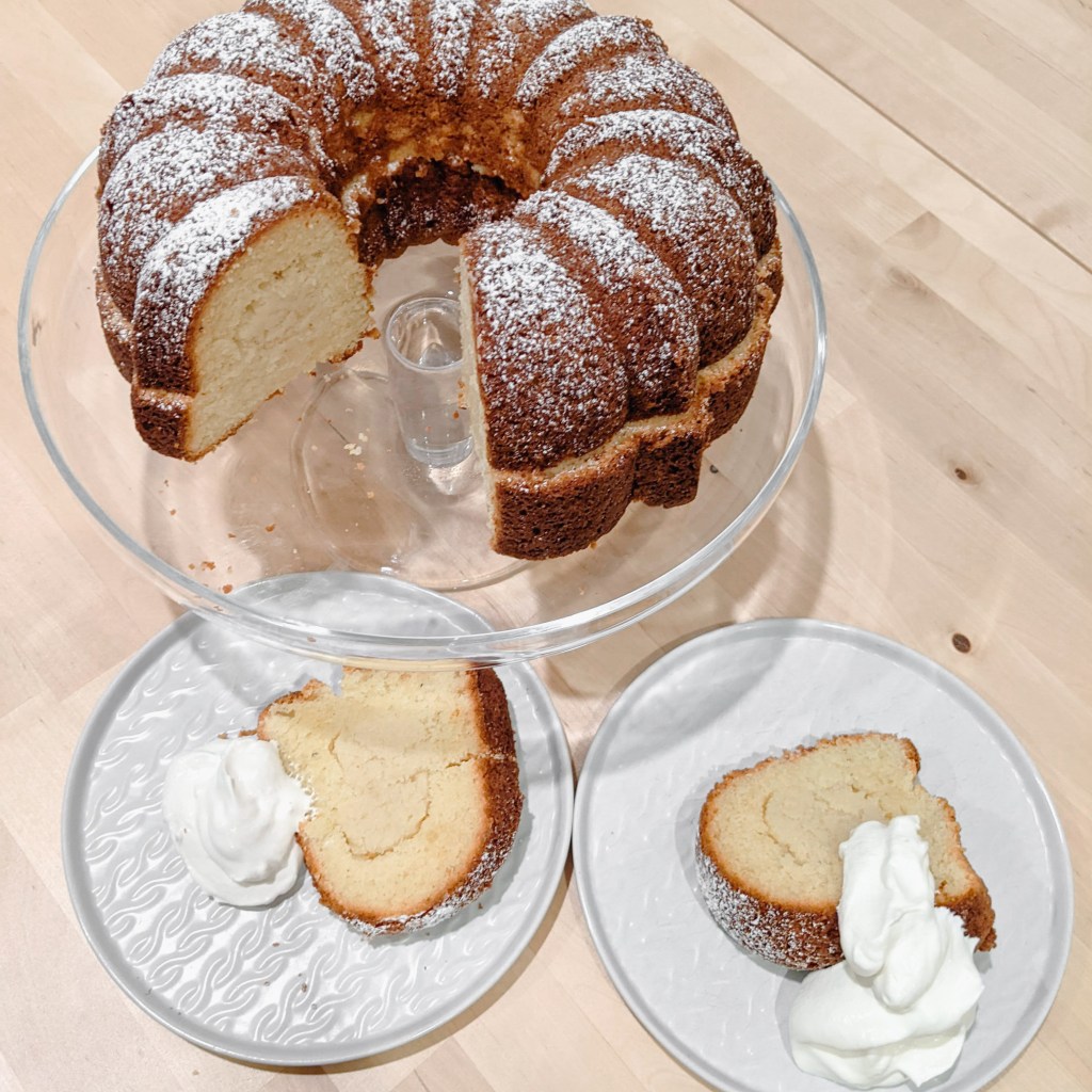 A bundt cake, dusted with powdered sugar and filled with almond paste, with two slices removed and plated with whipped cream.